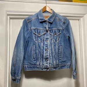 May include: A light blue denim jacket with a classic design. The jacket features a button-up front, two chest pockets, and two side pockets. The jacket is hanging on a wooden hanger against a white door.