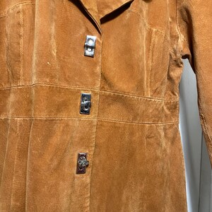 May include: A close-up of a tan suede coat with three silver metal clasps. The coat has a collar and a tailored fit. The suede has a soft, textured appearance. The coat appears to be vintage.