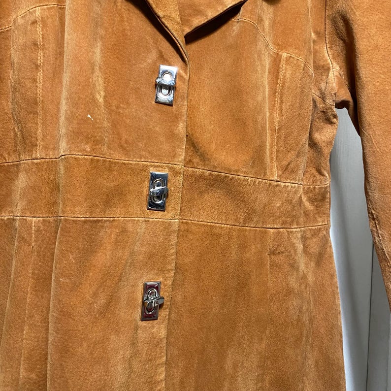 May include: A close-up of a tan suede coat with three silver metal clasps. The coat has a collar and a tailored fit. The suede has a soft, textured appearance. The coat appears to be vintage.
