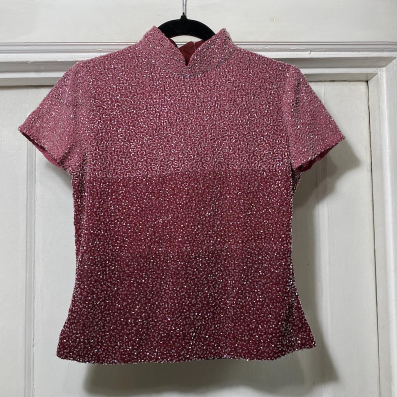 May include: A short-sleeved, burgundy top with a mandarin collar. The top is covered in small, shimmering sequins that create a gradient effect, transitioning from a lighter shade at the top to a deeper burgundy at the bottom. The garment hangs on a black hanger.