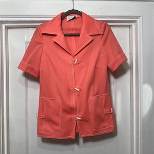 May include: A coral pink short-sleeved button-up shirt with a collar and two pockets. The shirt has white buttons and a white tag with the letter "D" on it.