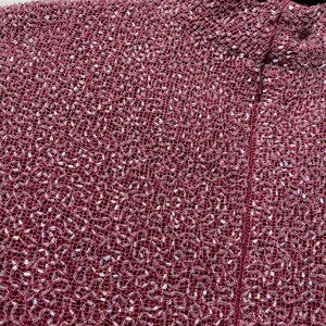 May include: Close-up of a textured, burgundy fabric densely covered with small, iridescent beads. The beads catch the light, creating a shimmering effect. The fabric appears to be a woven material, possibly for a garment.