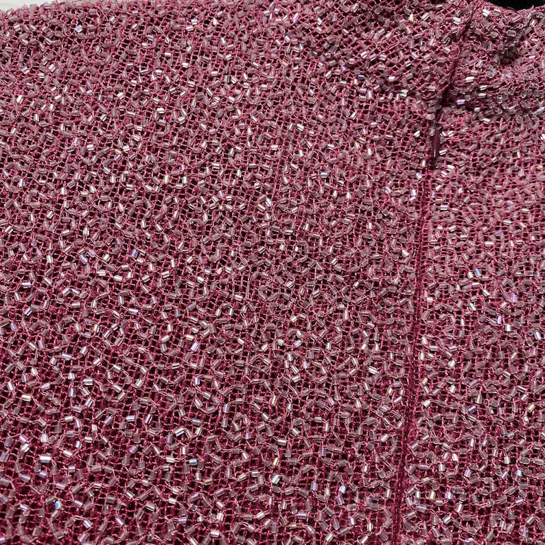 May include: Close-up of a textured, burgundy fabric densely covered with small, iridescent beads. The beads catch the light, creating a shimmering effect. The fabric appears to be a woven material, possibly for a garment.