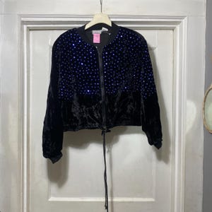 Puede incluir: Chaqueta bomber de terciopelo negro con cintura ajustable con cordón y cremallera frontal. La parte superior de la chaqueta está adornada con lentejuelas azules. La chaqueta está colgada en una percha.