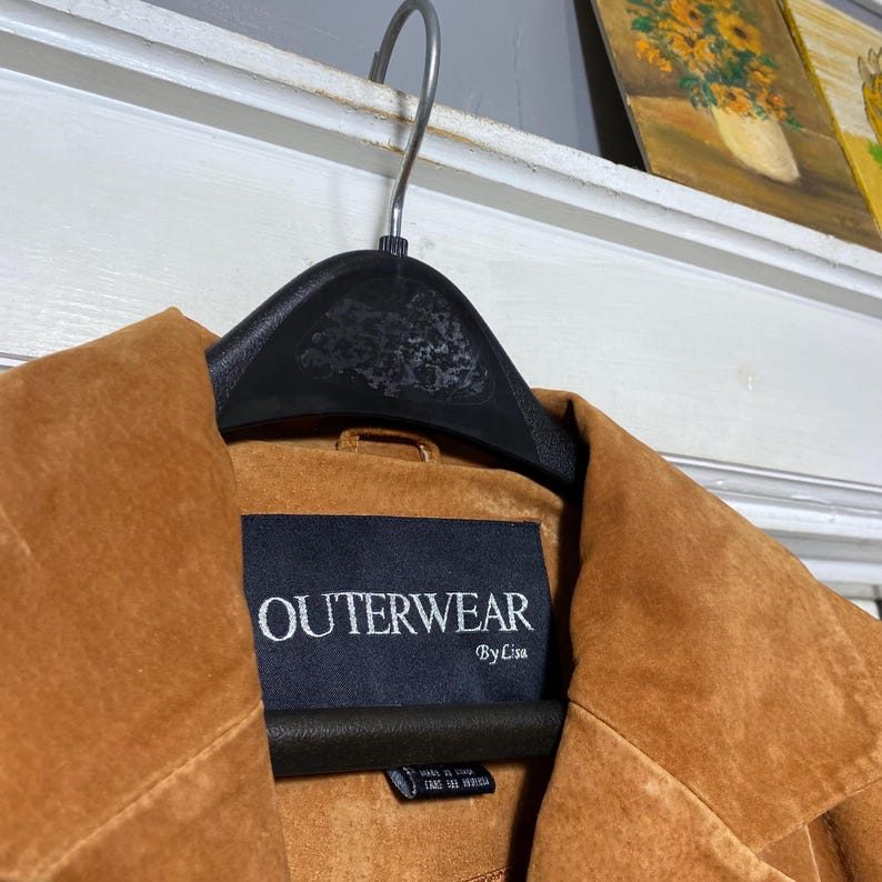 May include: A close-up of a brown suede jacket hanging on a black hanger. The jacket has a label that reads "OUTERWEAR By Lisa". The jacket is a warm, earthy tone, and the texture of the suede is visible.