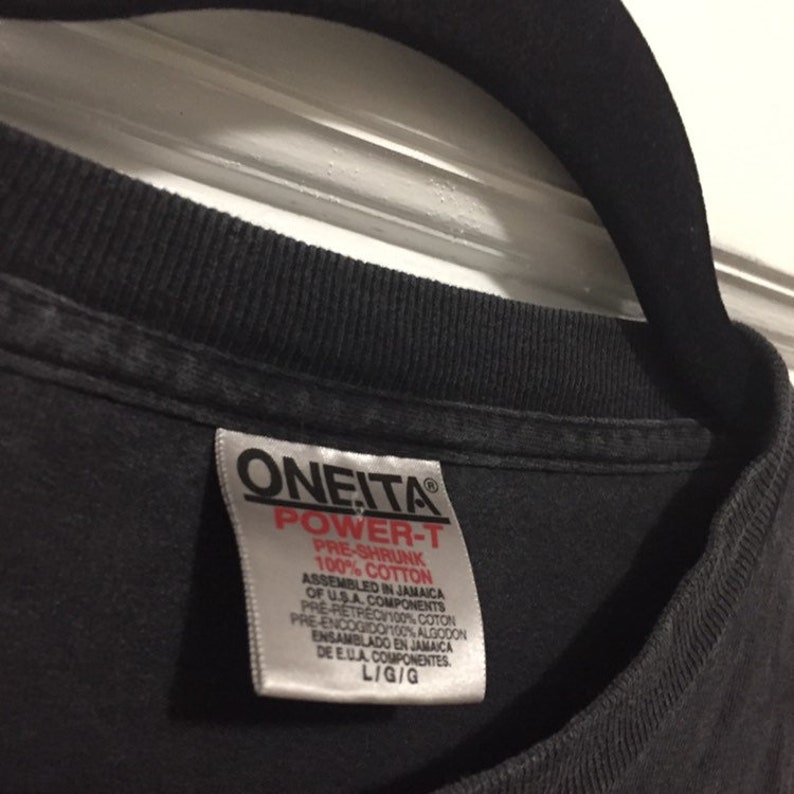 K&ouml;nnte beinhalten: Schwarzes T-Shirt mit einem wei&szlig;en Etikett, auf dem "Oneita Power-T Pre-Shrunk 100% Cotton Assembled in Jamaica of U.S.A. Components Pre-Retrec 100% Coton Pre-Encogido/100% Algodon Ensamblado en Jamaica de E.U.A. Componentes L/G/G" steht.