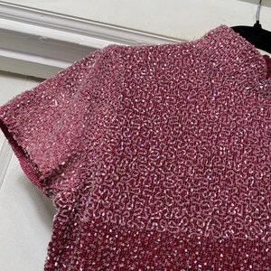 May include: Close-up of a short-sleeved, high-neck top in shades of pink and red. The fabric is densely beaded, creating a textured surface with a subtle shimmer. The garment's design features a gradient effect, transitioning from lighter pink at the sleeves to a deeper red.