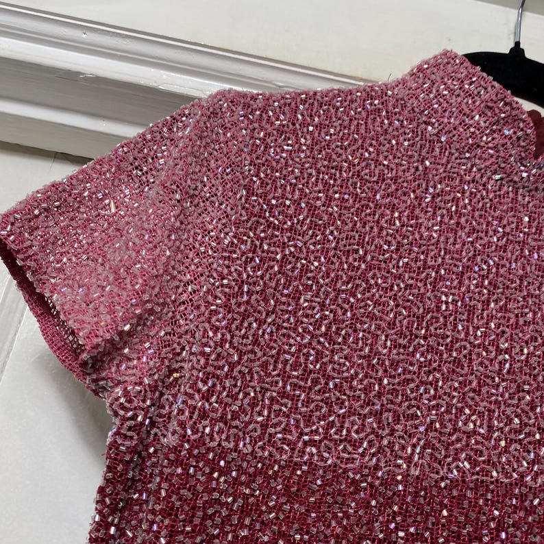 May include: Close-up of a short-sleeved, high-neck top in shades of pink and red. The fabric is densely beaded, creating a textured surface with a subtle shimmer. The garment's design features a gradient effect, transitioning from lighter pink at the sleeves to a deeper red.