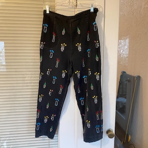 May include: Black denim pants with colorful embroidered floral designs. The embroidery features various flowers in vases, with red, yellow, blue, and white accents. The pants are hanging on a hanger.