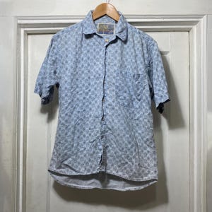 May include: Light blue short-sleeve button-down shirt with a subtle floral pattern. The shirt has a single chest pocket and is hanging on a wooden hanger. The fabric appears to be lightweight.