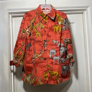 May include: A button-down shirt with a vibrant red background, featuring a safari animal print. The design includes giraffes, zebras, elephants, and lions. The long-sleeved shirt has black buttons and a collared neckline.