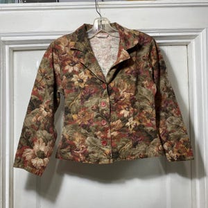 May include: A floral print blazer in shades of brown, green, and red. The jacket has a collared neckline, three buttons, and long sleeves. The fabric features a repeating pattern of stylized flowers and leaves.