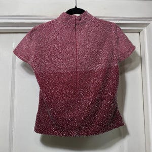 May include: A short-sleeved, burgundy-colored top with a high collar and a zipper down the front. The top is covered in small, sparkling embellishments, creating a textured, shimmering effect. The garment hangs from a black hanger.