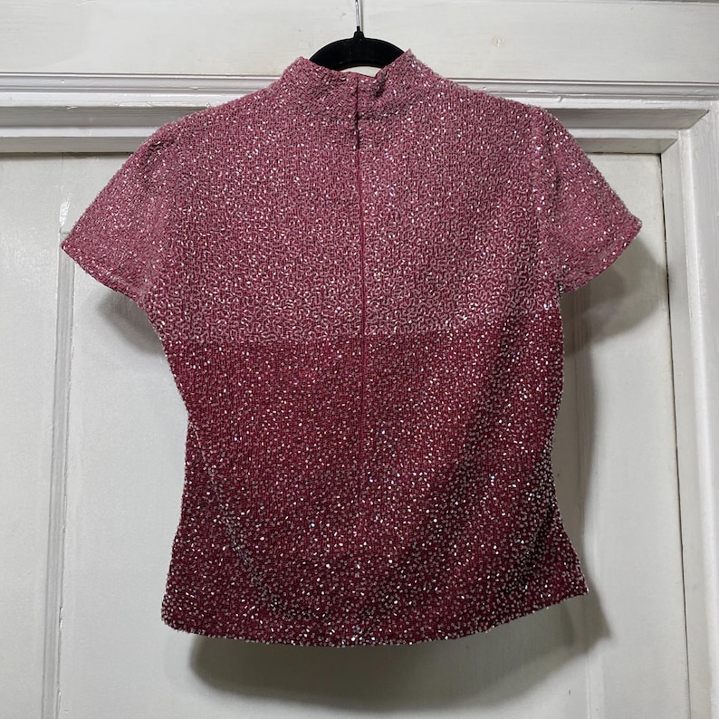 May include: A short-sleeved, burgundy-colored top with a high collar and a zipper down the front. The top is covered in small, sparkling embellishments, creating a textured, shimmering effect. The garment hangs from a black hanger.