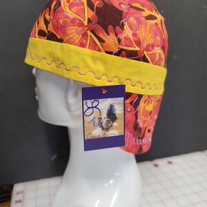 May include: A yellow and orange fabric welding cap with a butterfly pattern. The cap has a yellow band with a pink zig-zag stitch around the edge. The cap is on a white mannequin head. A purple tag with the text "JR Creations" is attached to the cap.