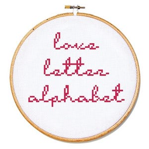 May include: A cross-stitch embroidery hoop with the words "love letter alphabet" stitched in red thread on white fabric. The wooden hoop is round and has a metal tightening mechanism.