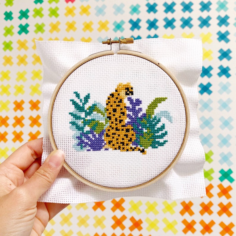 May include: A white embroidery hoop with a cross-stitch design of a yellow and black leopard sitting in a green and purple jungle scene.