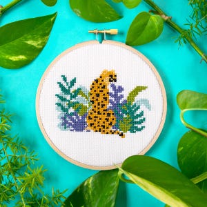 May include: A cross-stitch embroidery hoop with a white background featuring a yellow and black cheetah sitting in a jungle scene with green and purple leaves.