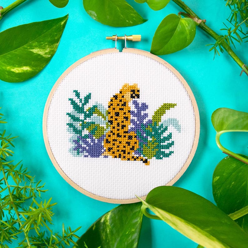 May include: A cross-stitch embroidery hoop with a white background featuring a yellow and black cheetah sitting in a jungle scene with green and purple leaves.