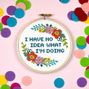 Puede incluir: Un aro de bordado blanco con un diseño floral y el texto "I have no idea what I'm doing". El aro está rodeado de círculos de fieltro de colores.