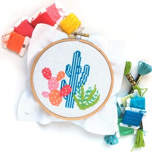 May include: A close-up of a completed cross-stitch project featuring a blue cactus with pink and orange cacti surrounding it. The project is framed in a wooden hoop and surrounded by colorful embroidery floss.