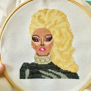 May include: Cross-stitch embroidery of a person with blonde hair wearing a black mock neck shirt. The person has brown skin and is wearing makeup.