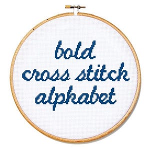 May include: A white cross-stitch hoop with the words "bold cross stitch alphabet" stitched in dark blue thread. The wooden hoop is round and has a brass clasp at the top.