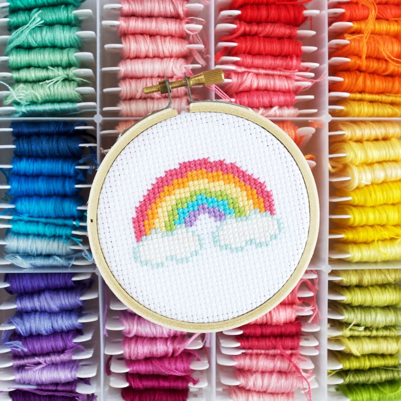 May include: A close-up of a rainbow cross-stitch embroidery project in a wooden hoop. The embroidery is surrounded by a variety of colorful embroidery floss spools.