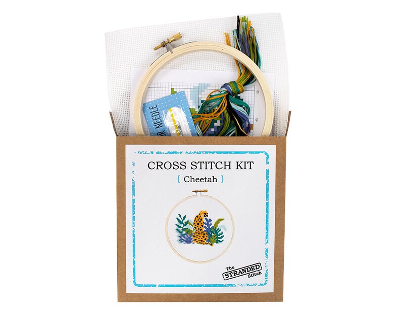May include: A cross stitch kit featuring a cheetah in a jungle setting. The kit includes a wooden embroidery hoop, Aida cloth, embroidery floss, and a needle. The box is brown with the text "CROSS STITCH KIT {Cheetah} The STRANDED Stitch"