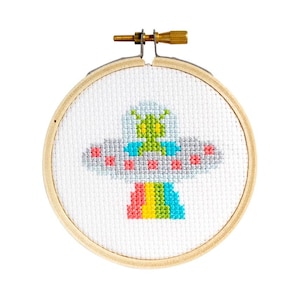 Puede incluir: Un bastidor de bordado de punto de cruz con un diseño de OVNI colorido y un extraterrestre verde. El OVNI tiene detalles en rojo, gris y azul, y una parte inferior de arcoíris. El bordado está en un aro de madera.