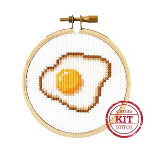 May include: A cross stitch kit featuring a simple design of a fried egg. The kit includes a wooden embroidery hoop, white Aida cloth, and embroidery floss. The kit is labeled "Cross Stitch Kit".
