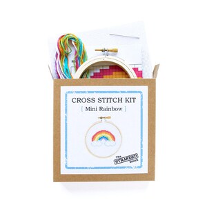May include: A cross stitch kit with a brown cardboard box, a wooden embroidery hoop, and colorful embroidery floss. The kit includes a pattern for a mini rainbow with clouds. The box has the text "CROSS STITCH KIT | Mini Rainbow | The STRANDED Stitch"
