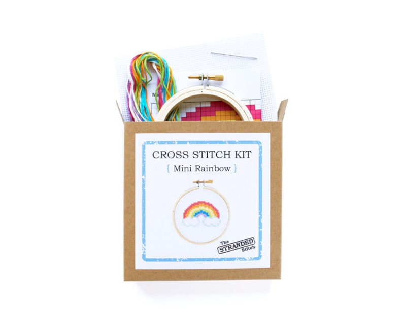 May include: A cross stitch kit with a brown cardboard box, a wooden embroidery hoop, and colorful embroidery floss. The kit includes a pattern for a mini rainbow with clouds. The box has the text "CROSS STITCH KIT | Mini Rainbow | The STRANDED Stitch"