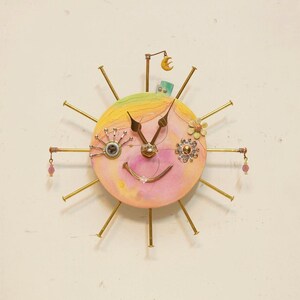 May include: A round wall clock with a pastel yellow and pink face featuring a smiling expression. The clock is surrounded by gold-toned nails acting as hour markers, with decorative elements including a small moon, a flower, and pink beads.