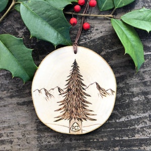 May include: A wood-burned ornament featuring a pine tree and mountains with the text "PCT" at the base of the tree.