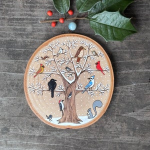 Peut inclure: Une plaque ronde en bois avec une image peinte d'un arbre avec des oiseaux et un écureuil. L'arbre est brun avec des points blancs ressemblant à de la neige. Les oiseaux sont rouges, bleus, noirs et blancs. L'écureuil est gris. La plaque est de couleur brun clair.
