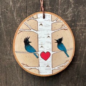 May include: A wooden ornament featuring a white birch tree with two blue birds perched on branches. A red heart is painted on the tree trunk.