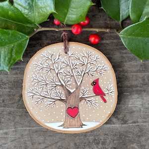 Peut inclure: Une décoration de Noël en bois avec un arbre peint en blanc, un cardinal rouge et un cœur rouge. La décoration est ornée de points blancs pour ressembler à la neige.