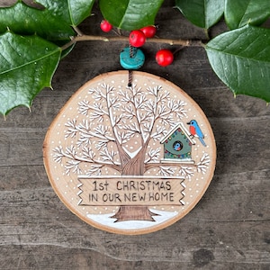 Peut inclure: Une décoration ronde en bois avec un arbre peint en blanc, un nichoir avec deux oiseaux bleus et le texte "1er Noël dans notre nouvelle maison".
