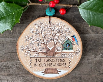 First Christmas in our new home wood slice ornament. Wooden sign on snowy tree with blue birds in a birdhouse. Handmade by Forage Workshop