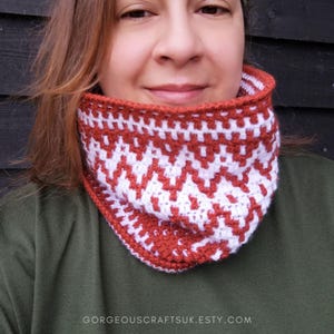 May include: A close-up of a woman wearing a crocheted cowl with a geometric pattern in white and rust orange. The cowl is folded over and the woman is looking at the camera.