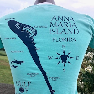 May include: A light blue t-shirt featuring a dark blue map of Anna Maria Island, Florida, with text including "Anna Maria Island Florida" and "Gulf of Mexico." The shirt also has a compass rose and dolphin graphics.
