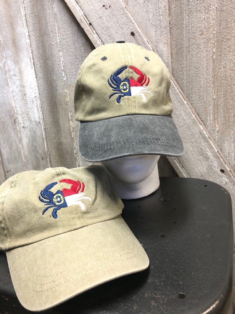 May include: Two khaki baseball caps with a crab design embroidered on the front. The crab is blue and red, with a white and blue design. One cap has a gray brim, and the other has a khaki brim. The caps are on a black surface.