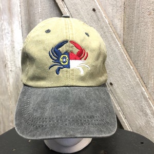 May include: A khaki baseball cap with a gray brim. The cap features an embroidered crab design in blue, red, white, and yellow, with the letters "N*C" inside a yellow circle. The hat is displayed on a black surface.