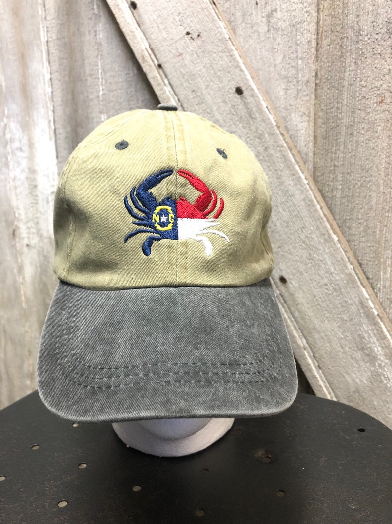 May include: A khaki baseball cap with a gray brim. The cap features an embroidered crab design in blue, red, white, and yellow, with the letters "N*C" inside a yellow circle. The hat is displayed on a black surface.