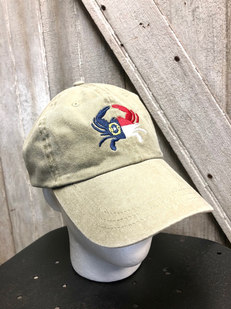 May include: A khaki-colored baseball cap with a crab design embroidered on the front. The crab is blue, red, white, and yellow. The hat is on a white mannequin head.