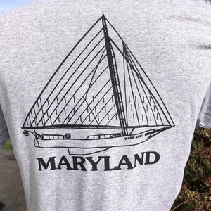 May include: A gray t-shirt featuring a black line drawing of a sailboat with the words "HARD SHELL" and "MARYLAND" printed below the boat. The shirt is made of a soft material.