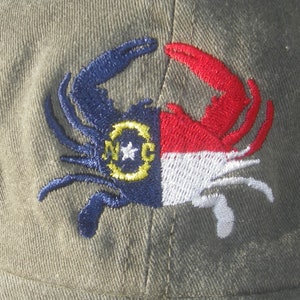 May include: Olive green baseball cap featuring an embroidered crab design. The crab is split, with one side in blue and the other in red and white, incorporating the North Carolina state flag. The crab's body includes the letters "NC" and a star.