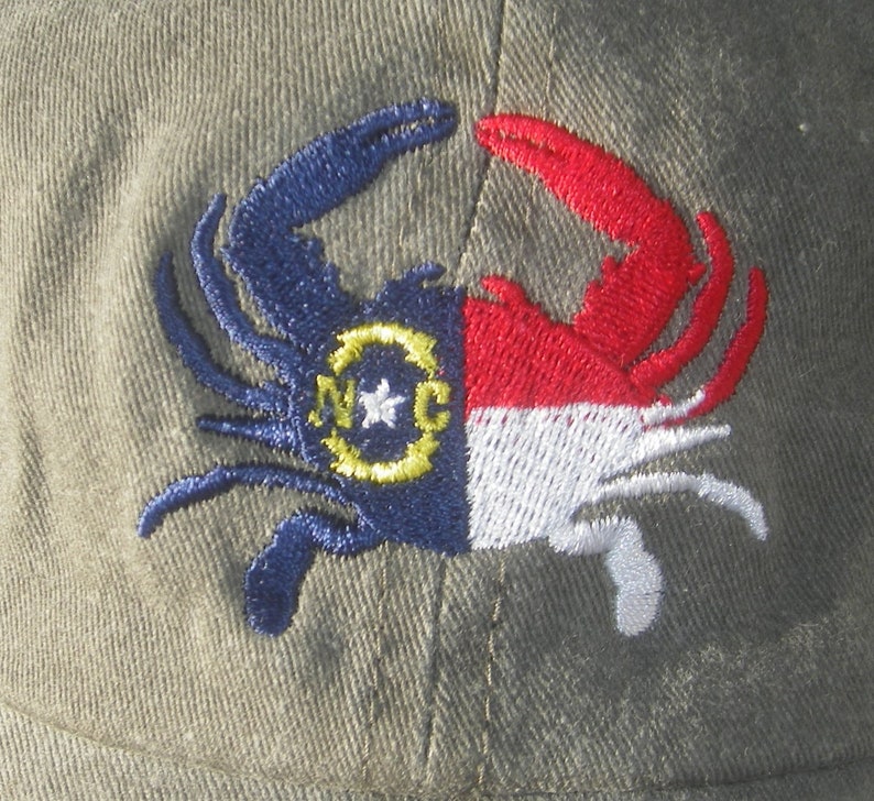 May include: Olive green baseball cap featuring an embroidered crab design. The crab is split, with one side in blue and the other in red and white, incorporating the North Carolina state flag. The crab's body includes the letters "NC" and a star.