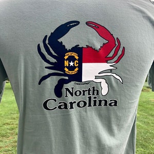 May include: Sage green t-shirt featuring a crab design with the North Carolina state flag colors. The crab's claws are navy and red, with the state seal in the center. The words "North Carolina" are printed below the crab.